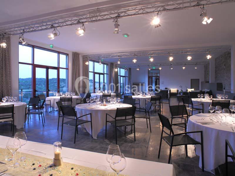 Salle de réception lumineuse avec tables rondes dressées, chaises noires et grandes baies vitrées donnant sur l'extérieur.