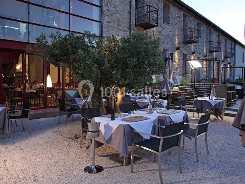 Terrasse d'un restaurant avec tables dressées, chaises modernes et un olivier au centre, devant un bâtiment en pierre.