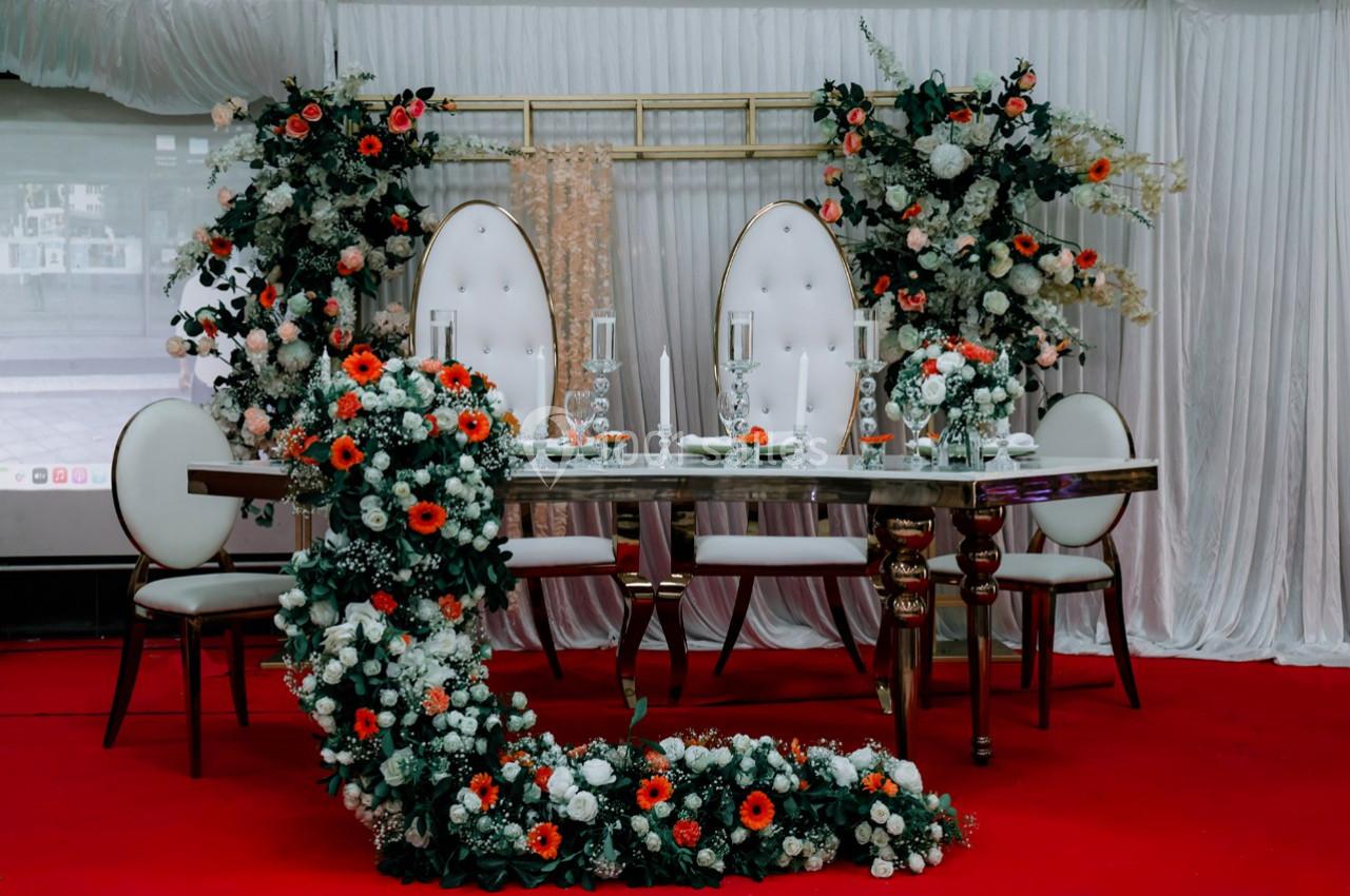 Table décorée pour un événement, entourée de chaises blanches et ornée de fleurs rouges, blanches et vertes.