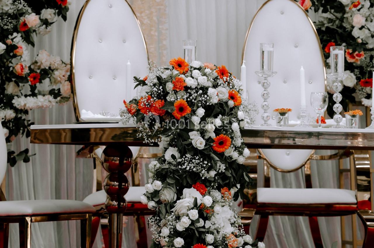 Table décorée pour un événement avec chaises blanches, fleurs orange et blanches, et chandeliers en verre.