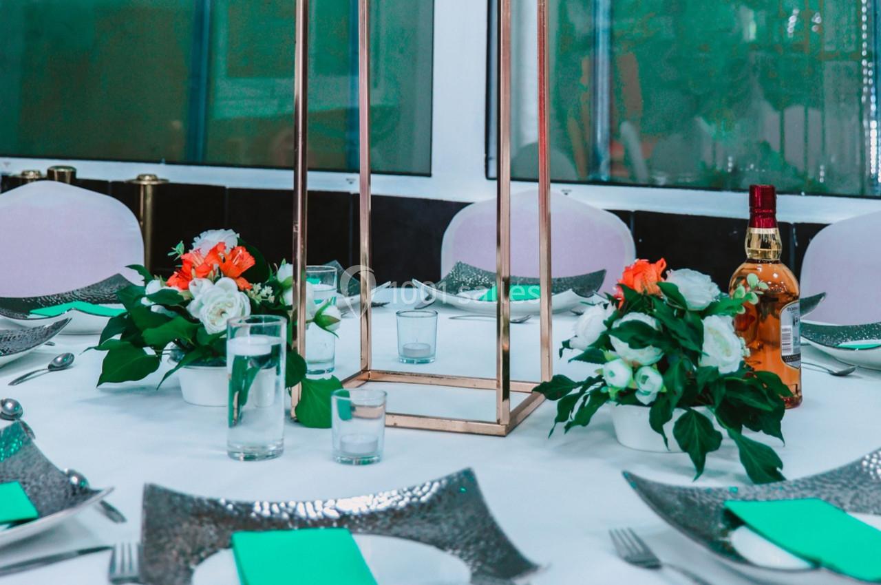 Table dressée avec nappes blanches, assiettes argentées, serviettes vertes, bouquets de fleurs et une bouteille en arrière…