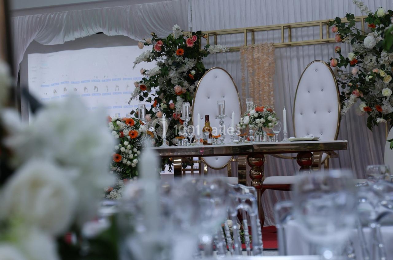 Table décorée pour un mariage avec chaises élégantes, fleurs blanches et orangées, et bougies sur fond de rideaux.