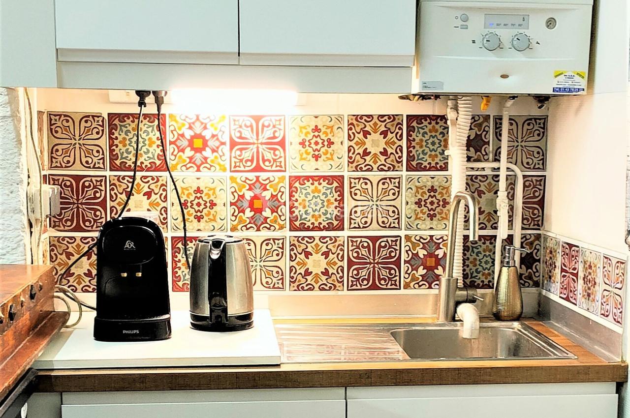Cuisine compacte avec évier, robinet, carrelage coloré, chauffe-eau mural, cafetière et grille-pain sur le plan de travail.