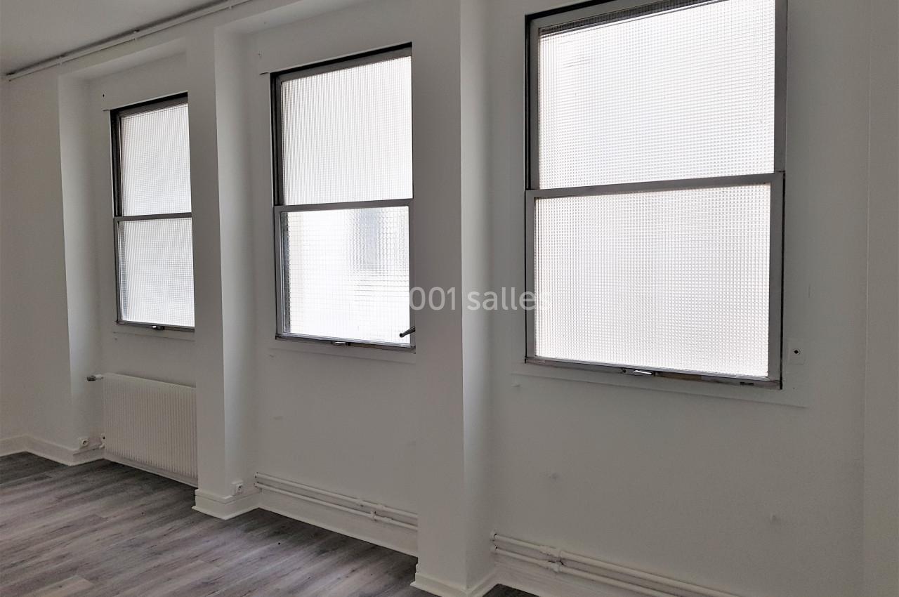 Trois fenêtres alignées dans une pièce lumineuse aux murs blancs et au sol en parquet gris.