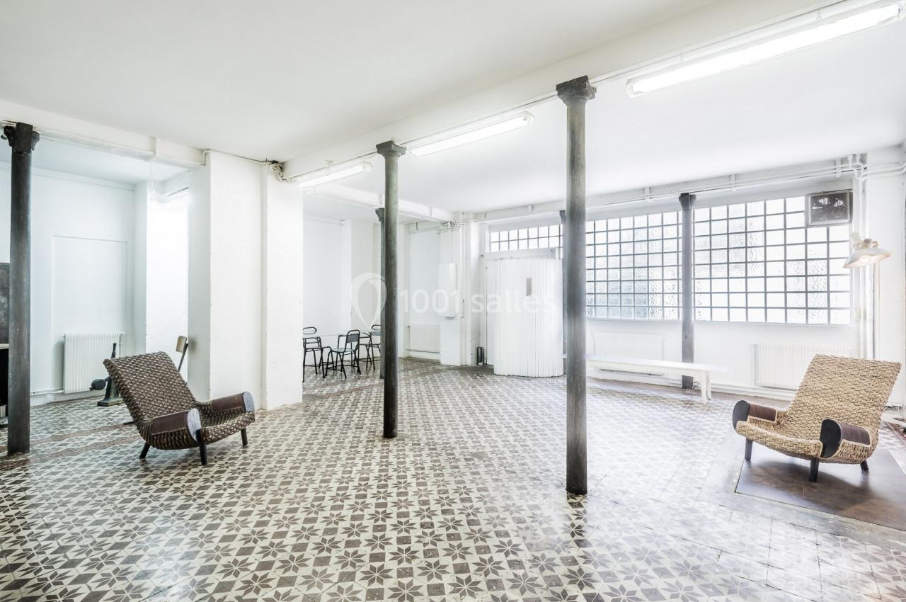 Salle lumineuse avec carrelage à motifs, colonnes métalliques, chaises et fauteuils en osier, grandes fenêtres en verre.