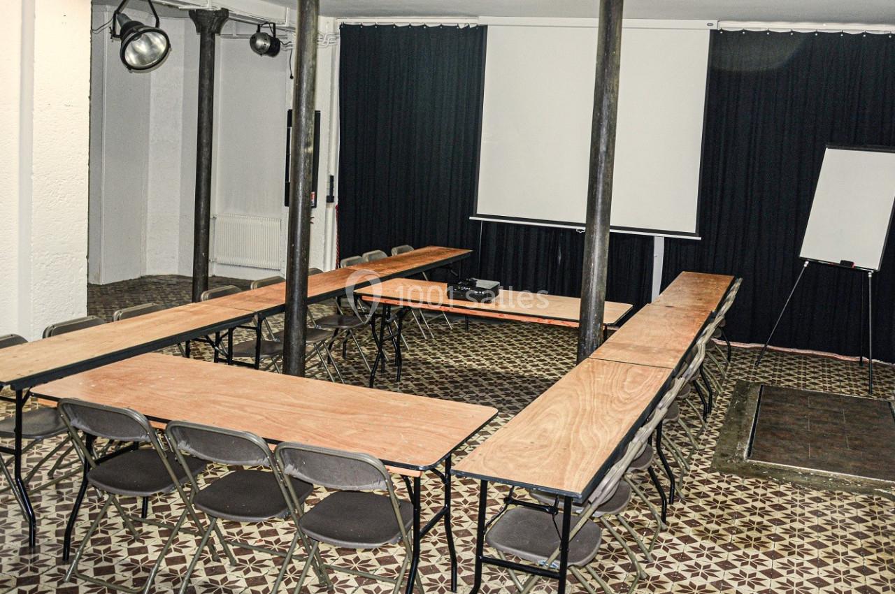 Salle de réunion avec tables disposées en U, chaises pliantes, écran de projection, paperboard et sol en carrelage.