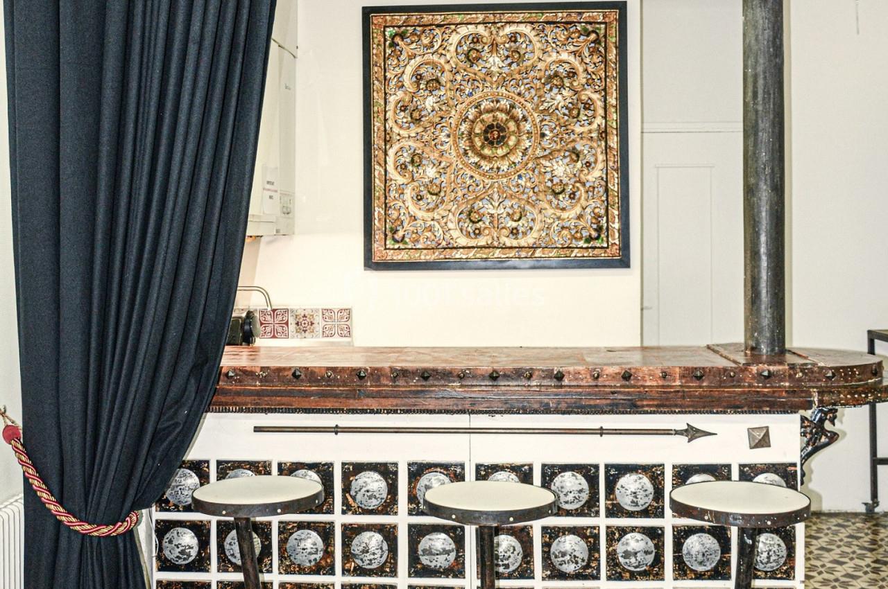 Comptoir en bois décoré, trois tabourets hauts, rideau noir à gauche et tableau ornemental au mur.