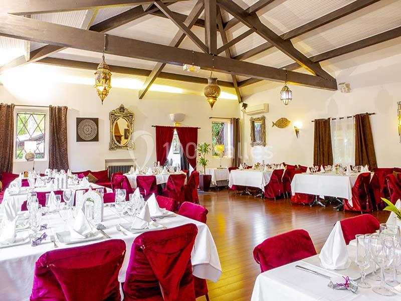 Salle de réception lumineuse avec tables dressées, nappes blanches, chaises rouges et décorations élégantes.