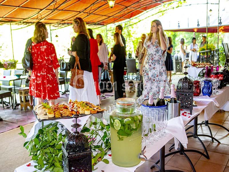 Des personnes discutent sous une terrasse couverte, près d'une table garnie de boissons et de plats variés.