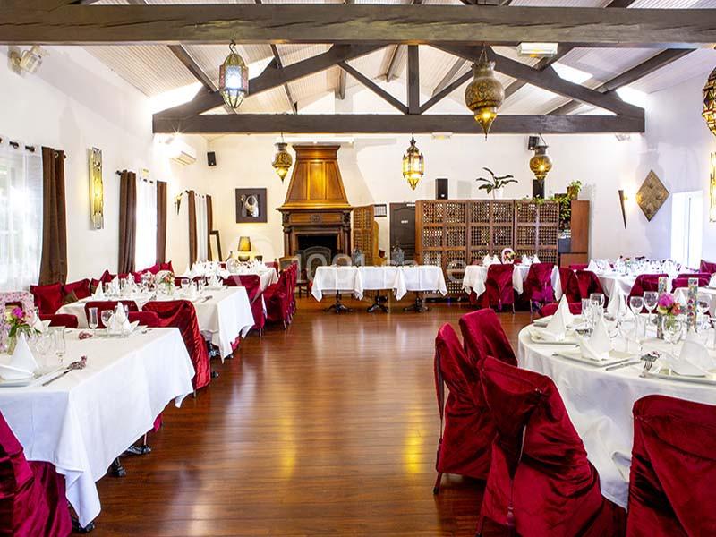 Salle de réception lumineuse avec tables dressées, chaises rouges, parquet en bois et décorations suspendues.