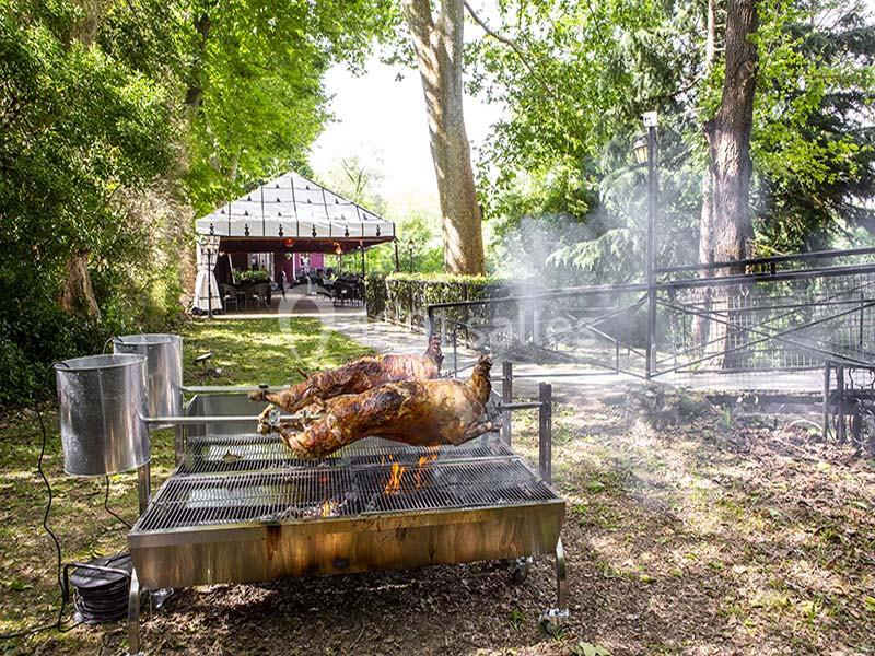 Deux agneaux rôtissent sur une broche au-dessus d'un feu en plein air, près d'une terrasse ombragée.