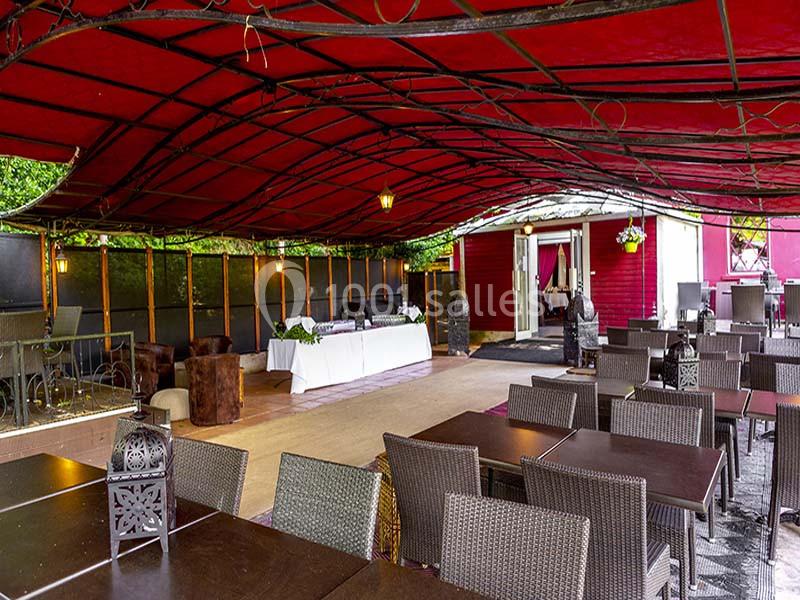 Terrasse couverte avec tables en bois, chaises en osier, buffet dressé et cabanon rouge en arrière-plan.