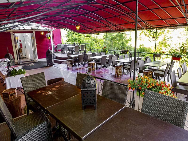 Terrasse couverte avec des tables en bois sombre, des chaises en osier et des plantes fleuries, entourée de verdure.