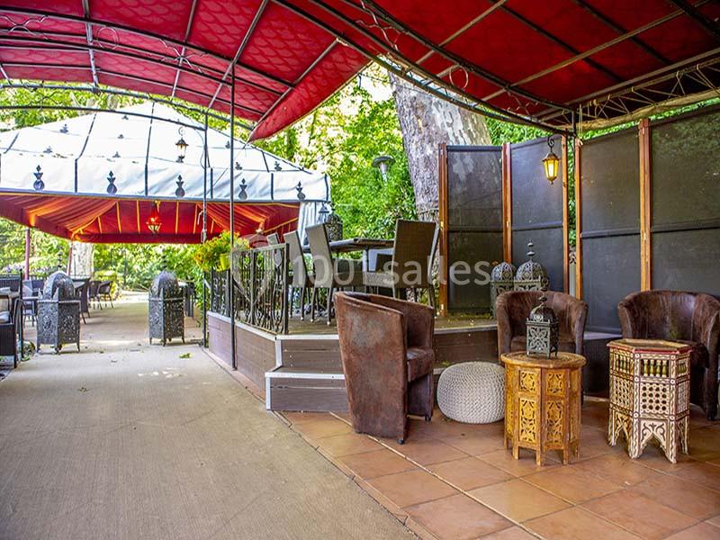 Terrasse extérieure aménagée avec fauteuils, tables en bois sculpté et pergolas rouges sous un cadre arboré.