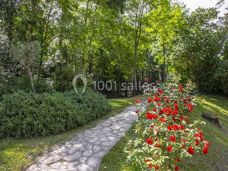 Allée en pierre bordée de buissons et de fleurs rouges, entourée d'arbres et de verdure sous un ciel dégagé.