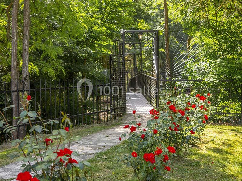 Allée bordée de rosiers rouges menant à une passerelle métallique entourée de verdure.