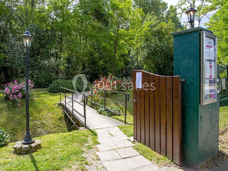 Entrée d'un parc avec portail en bois ouvert, chemin pavé, pont et végétation verdoyante en arrière-plan.