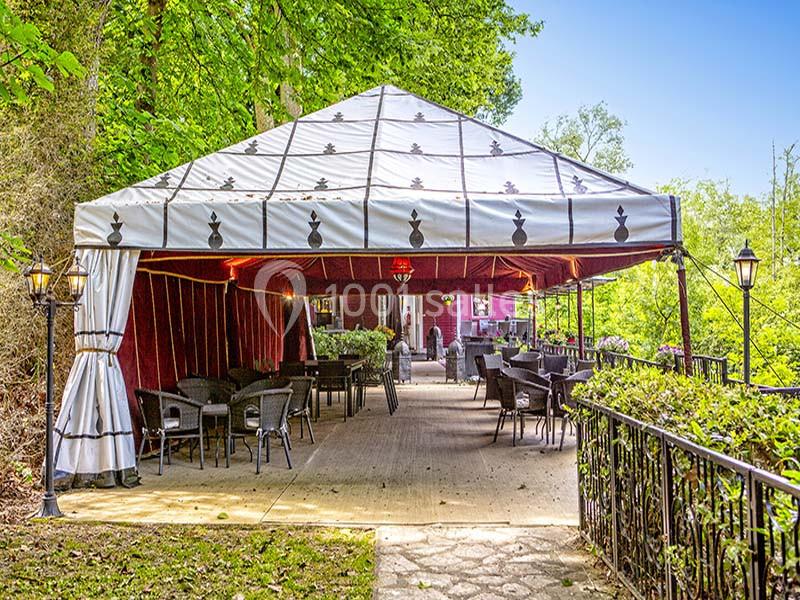 Tente extérieure aménagée avec des tables et des chaises, entourée de verdure et éclairée par des lampadaires.