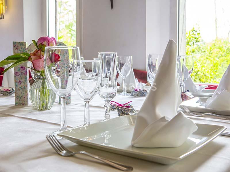 Table dressée avec serviettes pliées, verres et vaisselle, décorée de fleurs dans un vase près de fenêtres lumineuses.