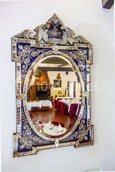Miroir orné de motifs dorés reflétant une salle de restaurant avec cheminée, tables dressées et chaises rouges.