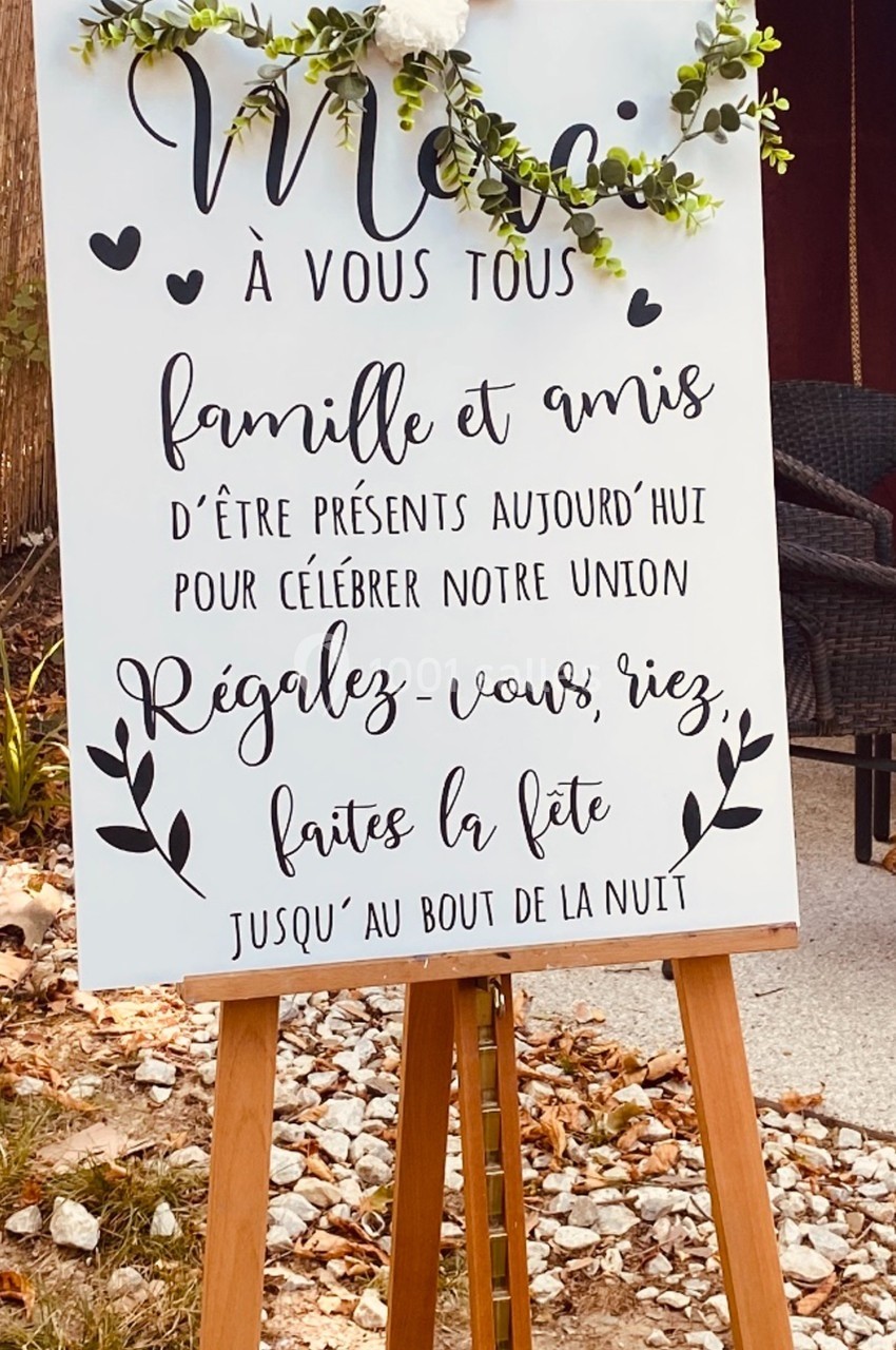 Panneau de mariage sur un chevalet, décoré de feuillage, invitant les invités à célébrer et faire la fête.