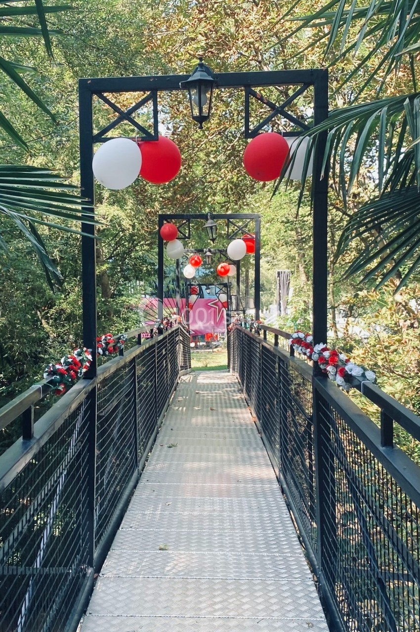 Passerelle décorée de ballons rouges et blancs, entourée de végétation dense et éclairée par des lampadaires.
