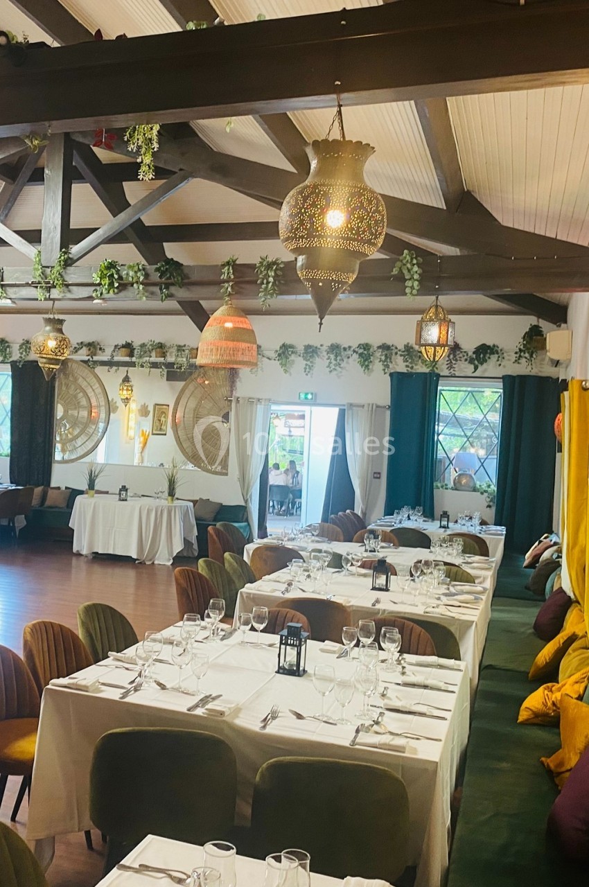 Salle de réception élégante avec tables dressées, décorations suspendues et lumière naturelle entrant par les fenêtres.