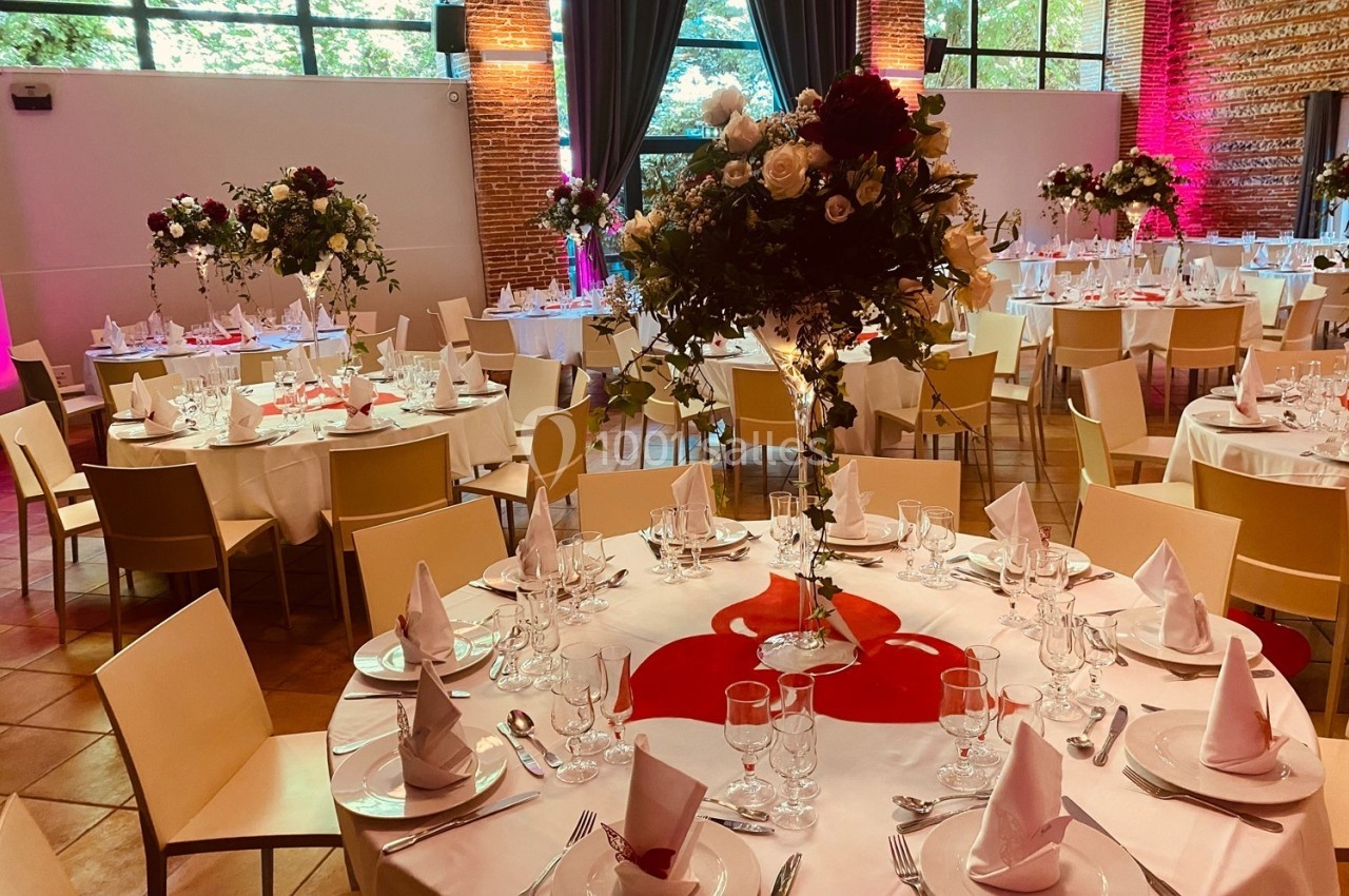 Salle de réception décorée avec des tables rondes, nappes blanches, centres de table fleuris et lumière naturelle.