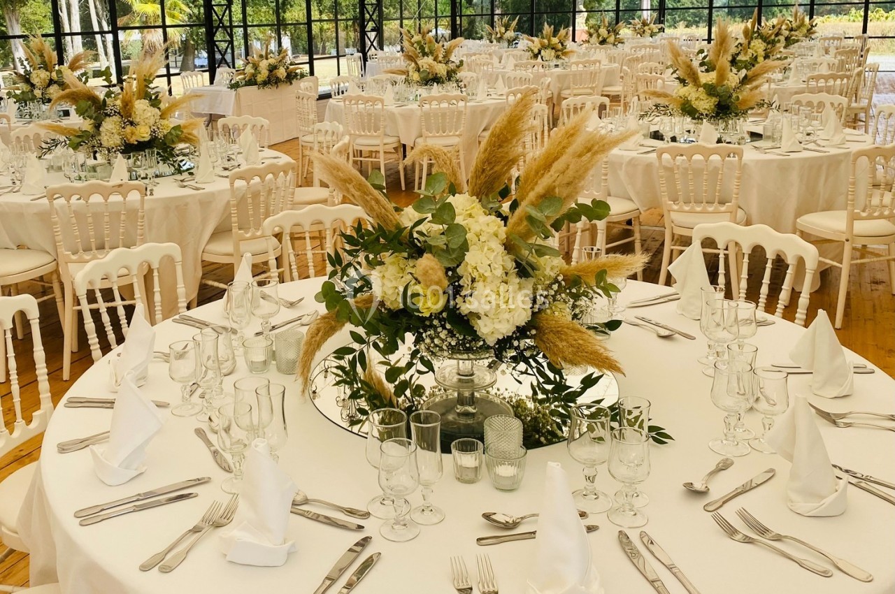Salle de réception décorée avec des tables rondes, nappes blanches, centres de table floraux et chaises blanches.