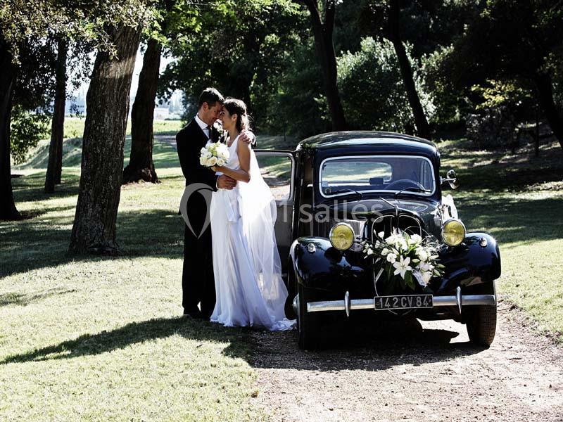 Location salle Piolenc (Vaucluse) - Château Beauchêne #15 Un couple de mariés pose près d'une voiture ancienne décorée de fleurs dans un parc verdoyant.