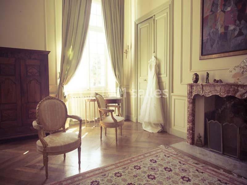 Location salle Piolenc (Vaucluse) - Château Beauchêne #12 Robe de mariée suspendue dans une pièce élégante avec meubles anciens, cheminée et grandes fenêtres.