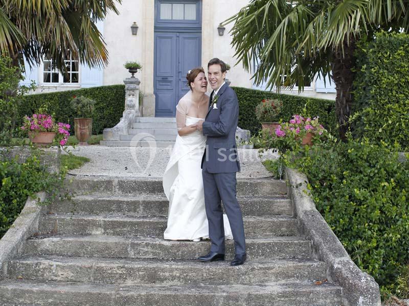Location salle Piolenc (Vaucluse) - Château Beauchêne #17 Un couple en tenue de mariage sourit sur les marches d'un escalier en pierre devant une maison entourée de végétation.