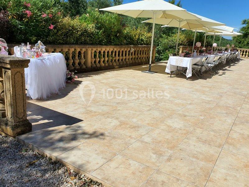 Terrasse ensoleillée avec tables décorées pour un événement, entourée de verdure et protégée par des parasols blancs.