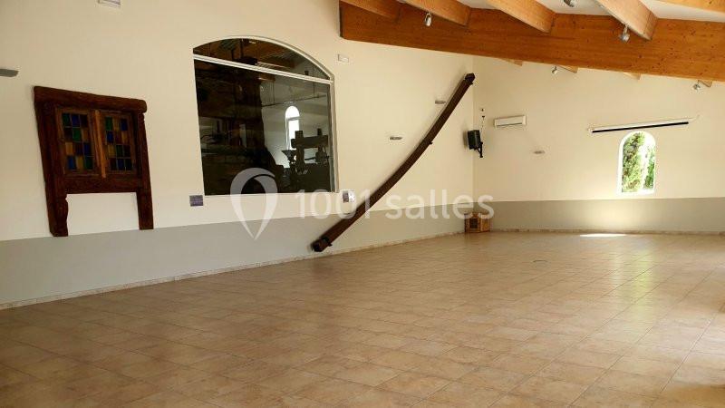 Salle vide avec sol carrelé, murs clairs, poutres apparentes au plafond et grandes fenêtres laissant entrer la lumière…