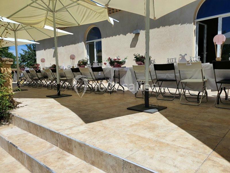 Terrasse ensoleillée avec tables dressées pour un repas, chaises alignées et parasols ouverts devant un bâtiment clair.