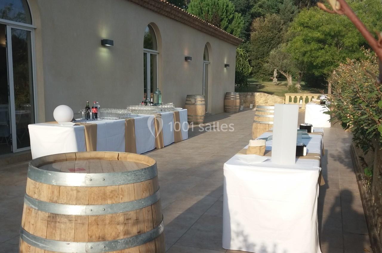 Terrasse ensoleillée avec tables dressées, tonneaux en bois et vue sur un jardin arboré.