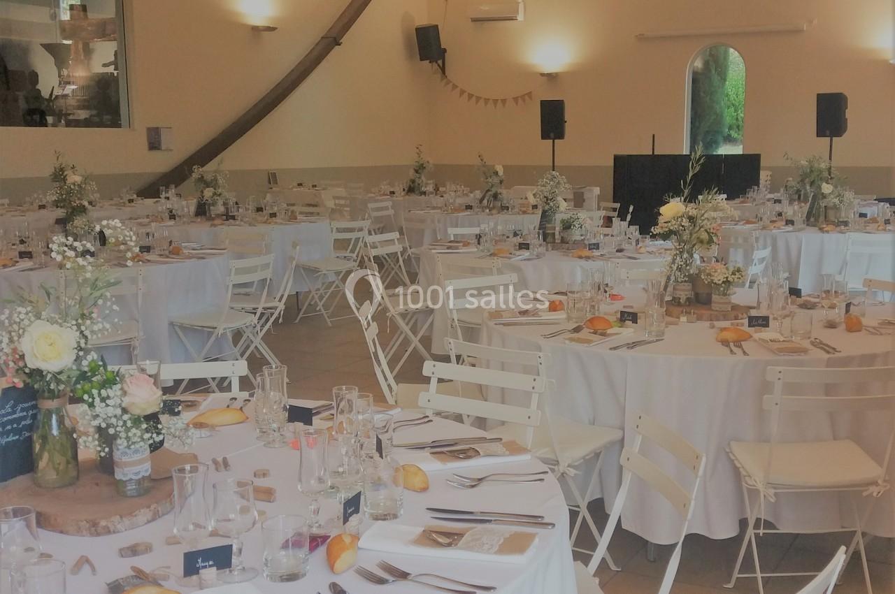 Salle de réception lumineuse avec tables rondes dressées, nappes blanches, chaises pliantes et décorations florales.