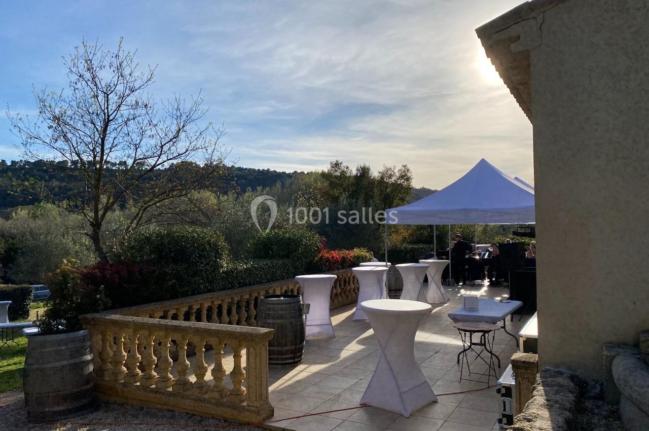 Terrasse ensoleillée avec tables hautes, tonnelle blanche et vue sur un paysage arboré en arrière-plan.
