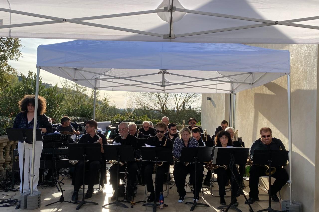 Un groupe de musiciens joue sous une tente blanche en extérieur, avec des pupitres et des instruments à vent.