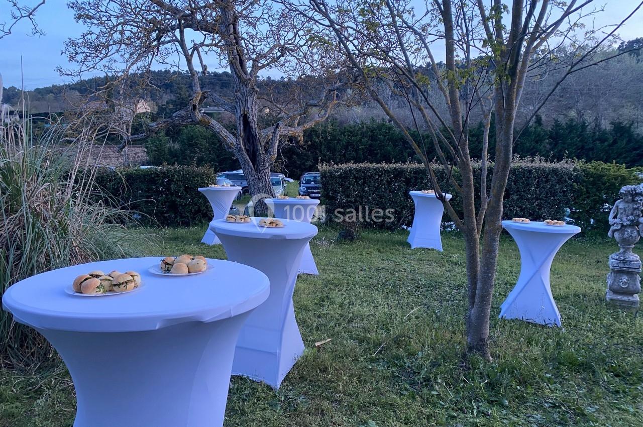 Tables hautes blanches dressées en extérieur avec des plateaux de petits sandwichs, entourées de végétation et d'arbres.