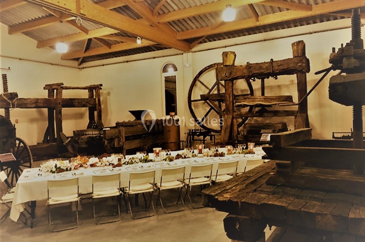Salle avec de grandes presses anciennes en bois et une longue table dressée pour un repas.