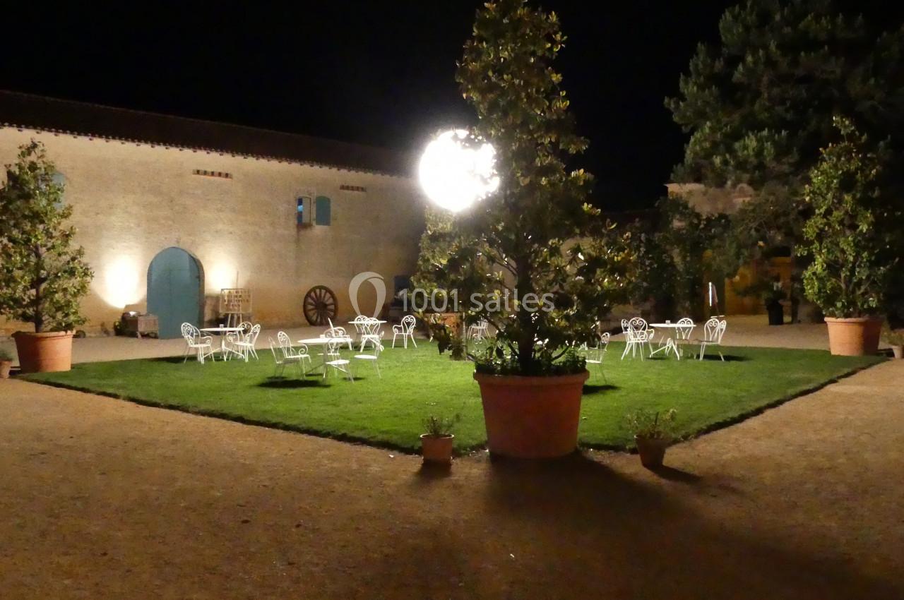 Cour extérieure éclairée la nuit, avec des tables et chaises blanches disposées autour d'une pelouse centrale.