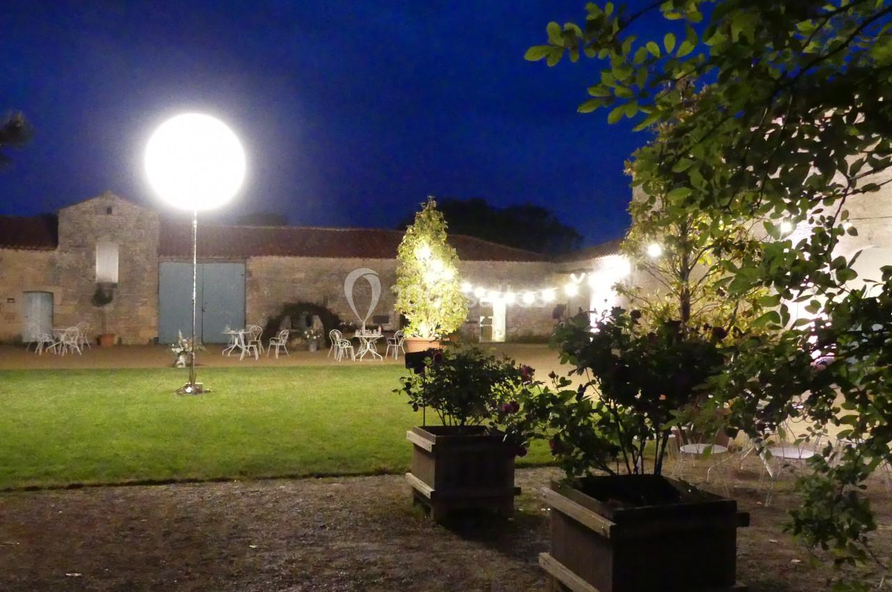 Cour extérieure éclairée de nuit avec des guirlandes lumineuses, des tables et des plantes en pot.
