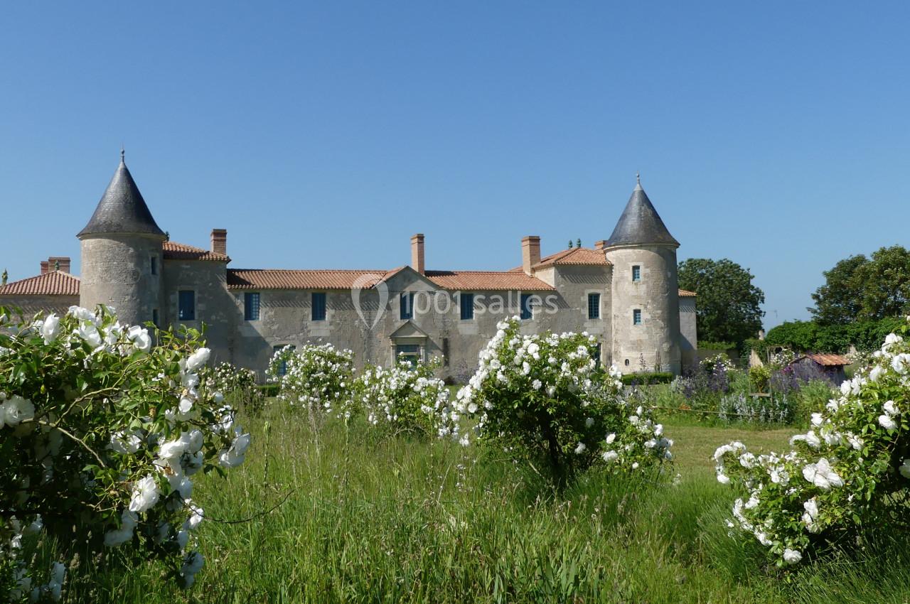 Château en pierre avec deux tours, entouré de rosiers blancs et d'un jardin verdoyant sous un ciel bleu.