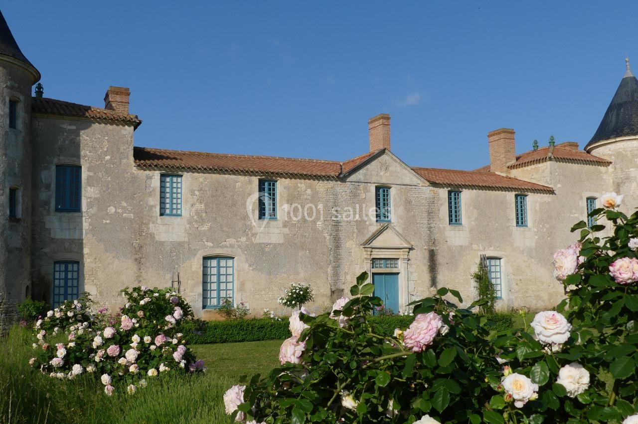 Façade d'un château ancien en pierre avec des tours, entouré de rosiers en fleurs sous un ciel bleu.