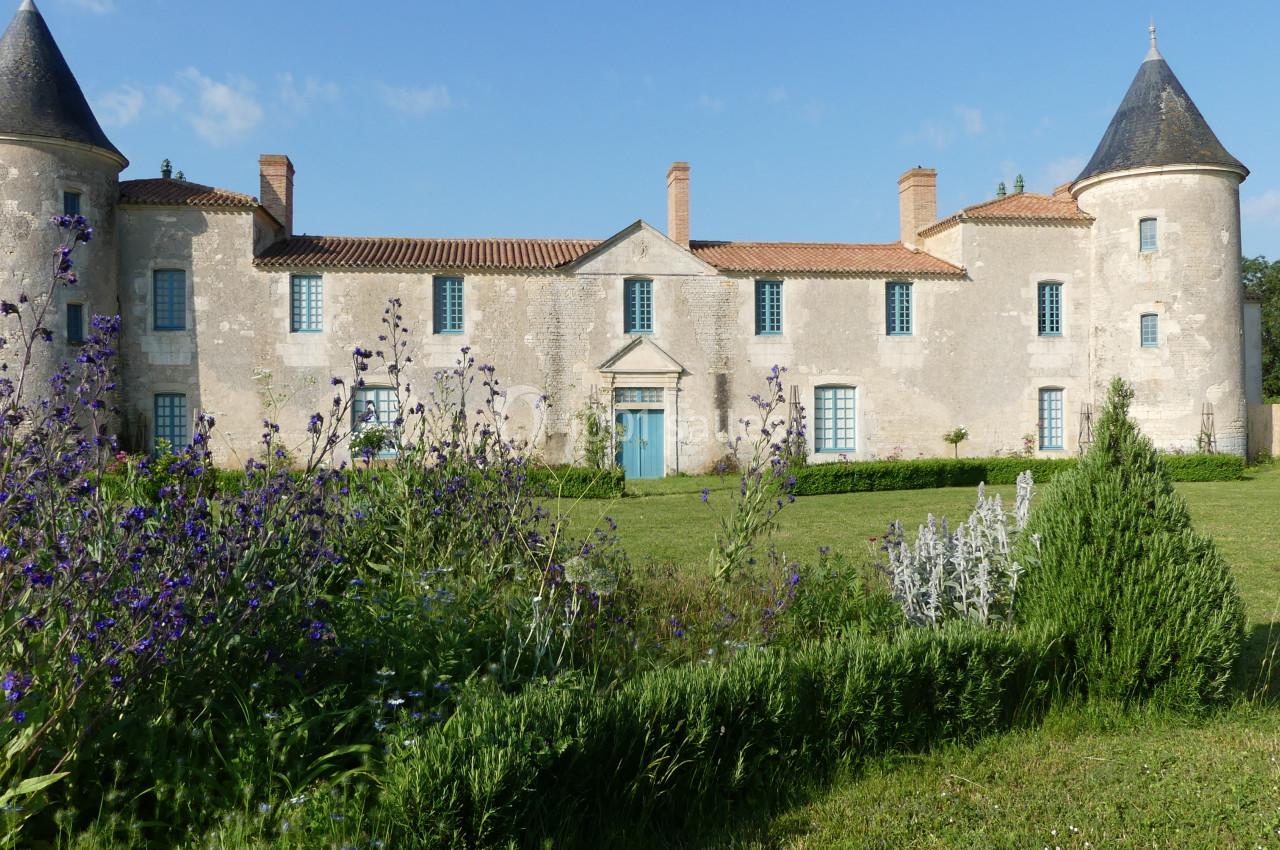Façade d'un château ancien avec deux tours rondes, entouré de jardins fleuris et d'une pelouse verte.