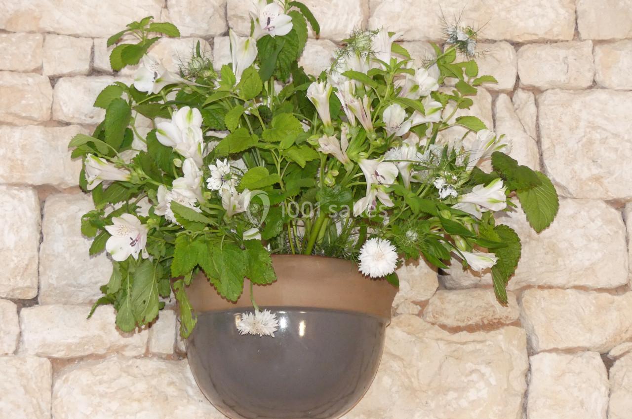 Pot mural en céramique contenant un bouquet de fleurs blanches et de feuillage vert, fixé sur un mur en pierre beige.