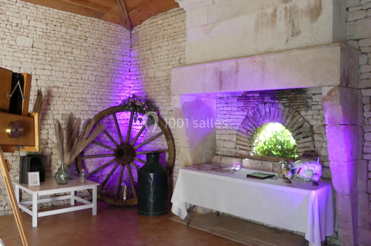 Salle en pierre avec une cheminée, une roue en bois décorative, une table blanche et un éclairage violet.