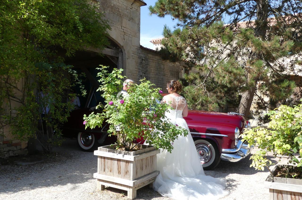 Une mariée en robe blanche se tient près d'une voiture rouge ancienne, entourée de verdure et de bâtiments en pierre.