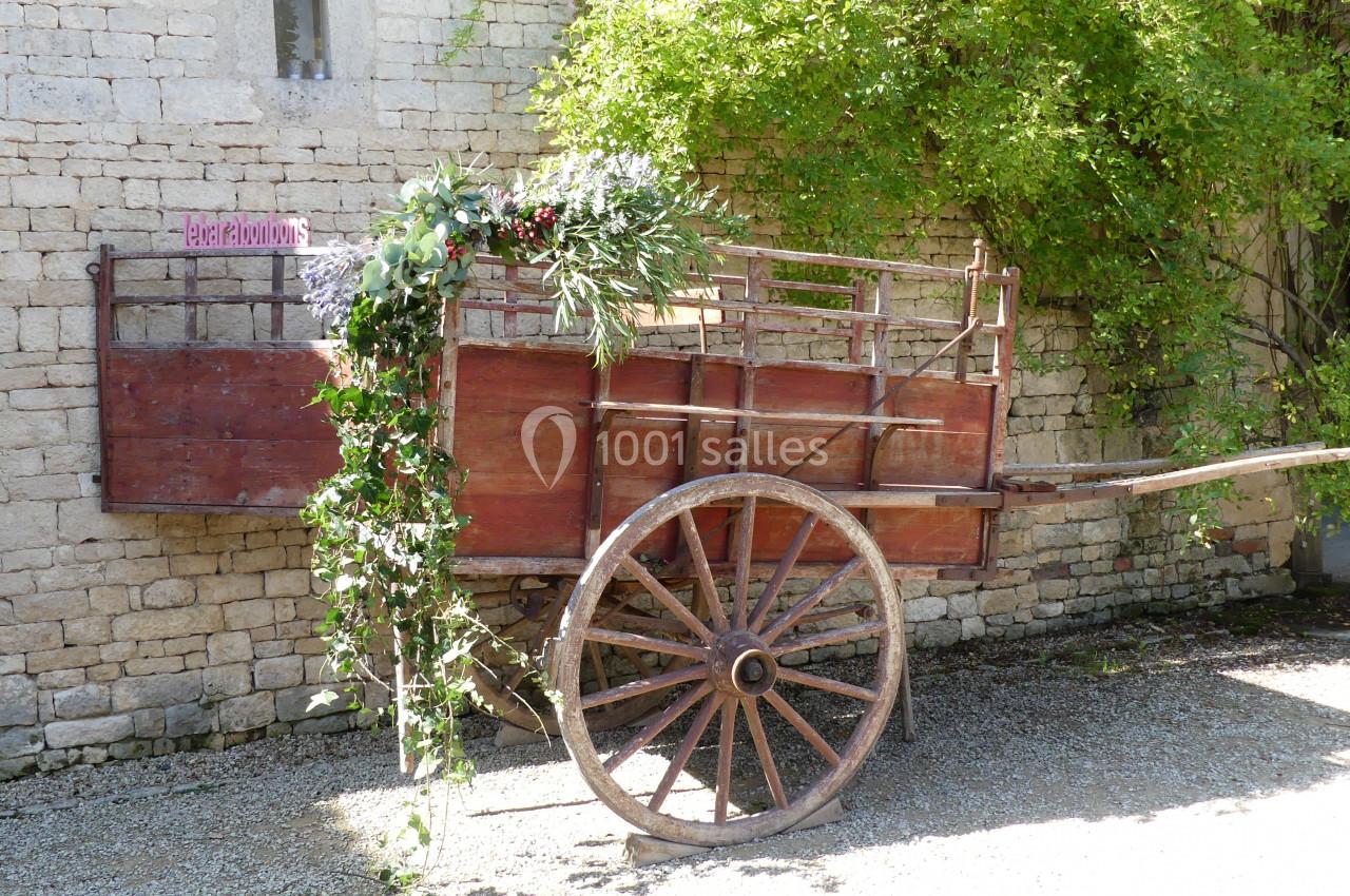 Charrette en bois décorée de fleurs et feuillages, adossée à un mur en pierre dans un espace extérieur ensoleillé.
